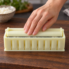 A hand presses down on the light yellow plastic sushi maker with a long, curved top piece, forming the roll.