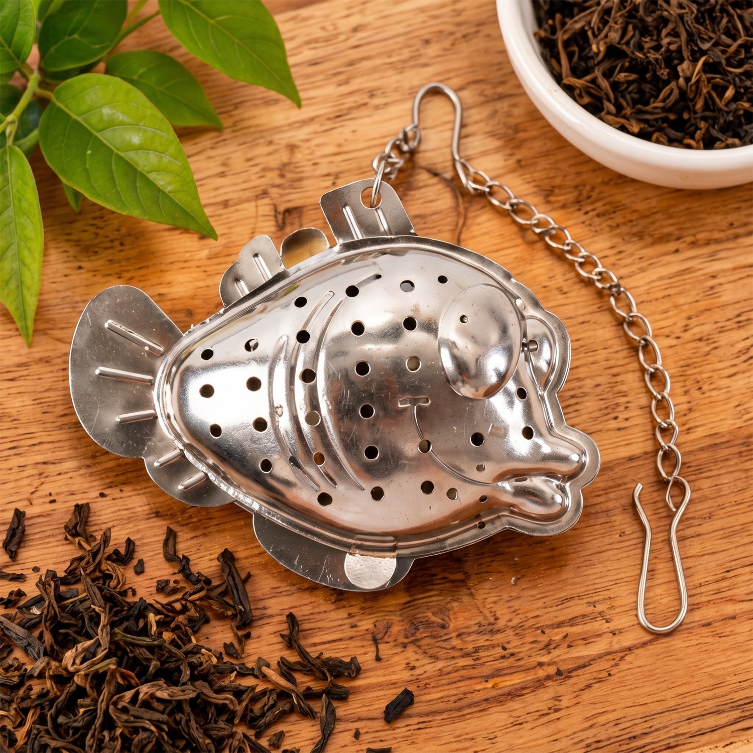 A closed silver fish-shaped tea infuser with its chain and hook, surrounded by loose dark tea leaves on a wooden surface.