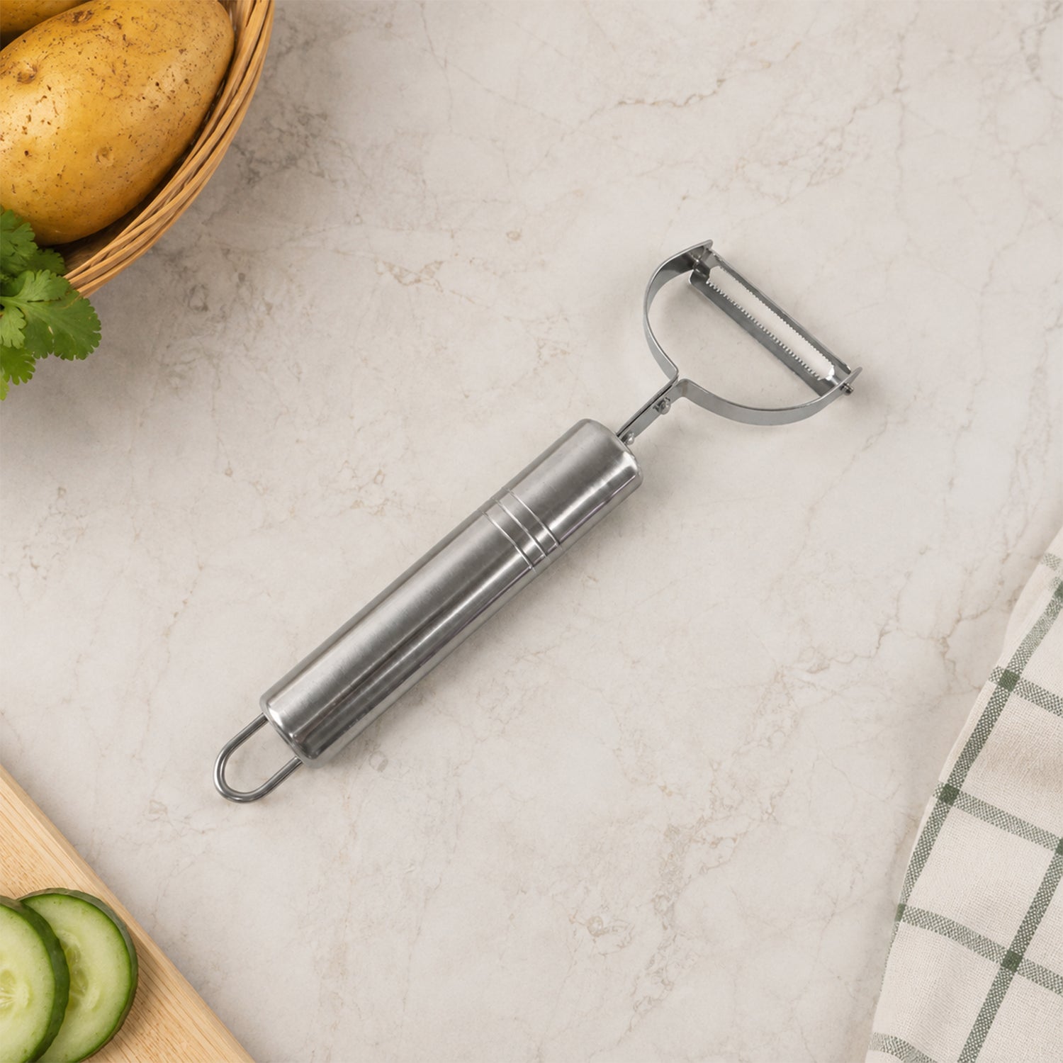 A stainless steel peeler on a light speckled surface next to a basket of potatoes, sliced cucumber, and a green plaid towel.