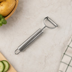 A stainless steel peeler on a light speckled surface next to a basket of potatoes, sliced cucumber, and a green plaid towel.