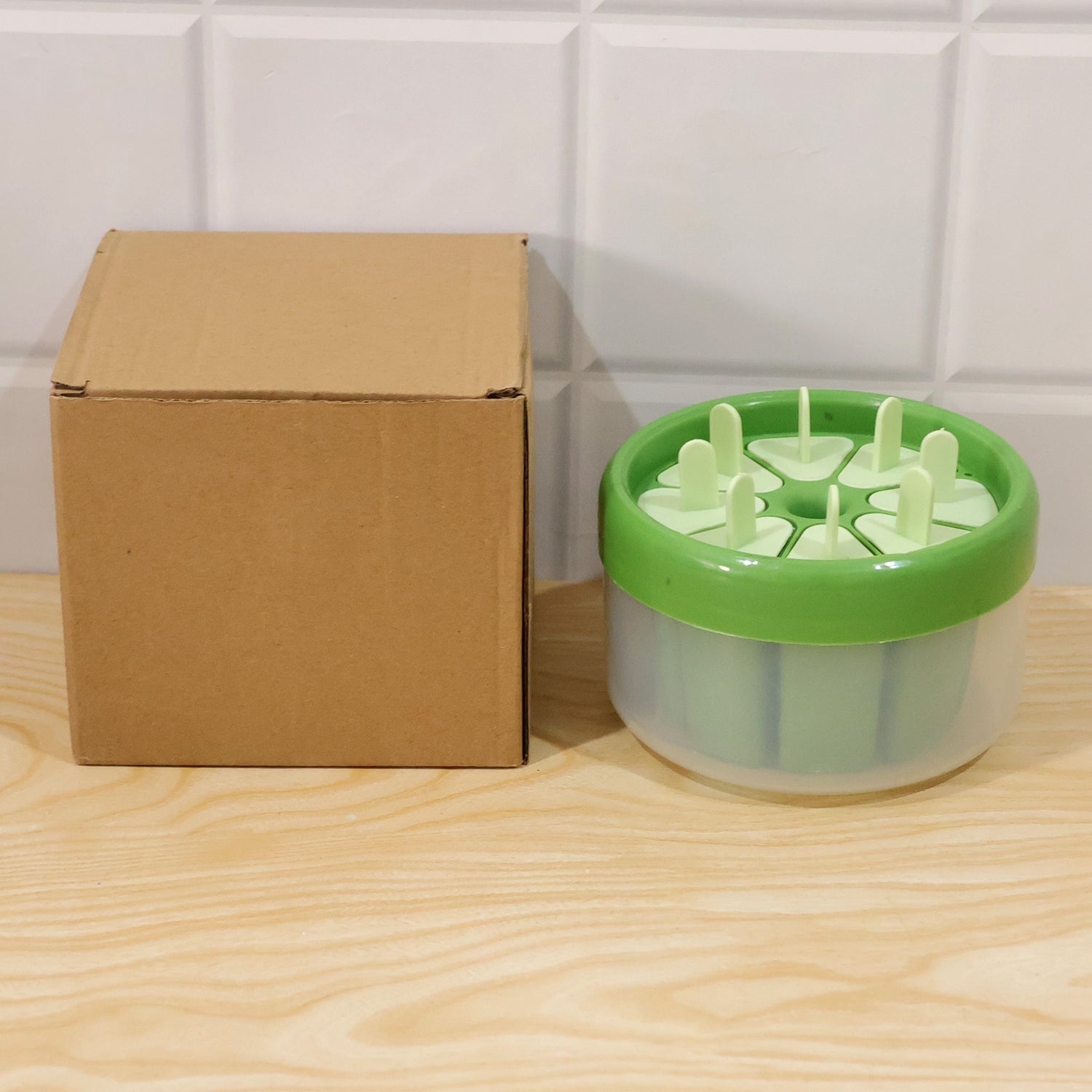 A green and white round plastic ice cream mold with its eight stick lids, next to a plain brown cardboard box on a wooden