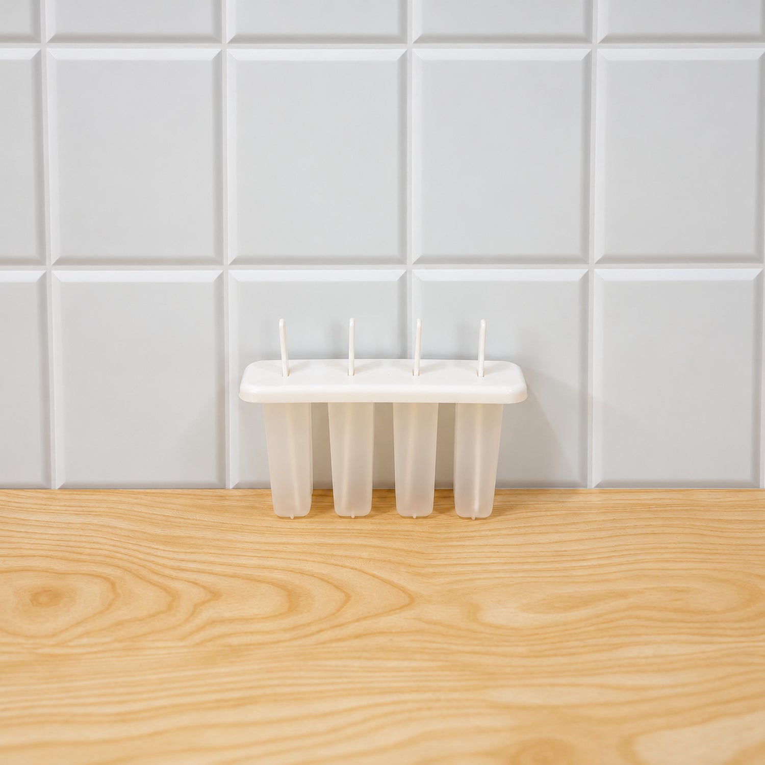 A white plastic popsicle mold with four cavities and sticks sits on a light wooden surface against a white tiled wall.