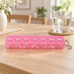 A pink plastic weekly pill box with seven compartments, each labeled with a Cyrillic day, filled with various pills on a