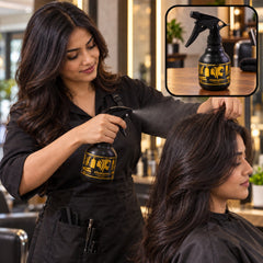 A stylist in a black apron sprays a client's long dark hair with a black and gold bottle in a salon setting.