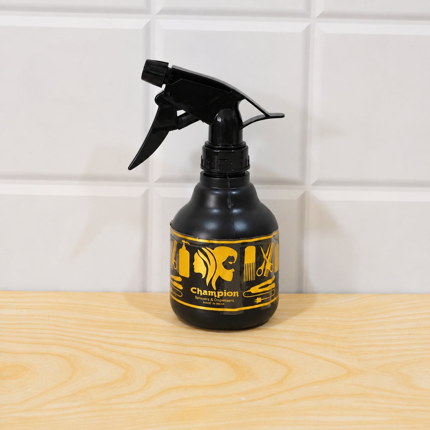 A black plastic spray bottle with a gold label rests on a light wooden surface against a white tiled wall.