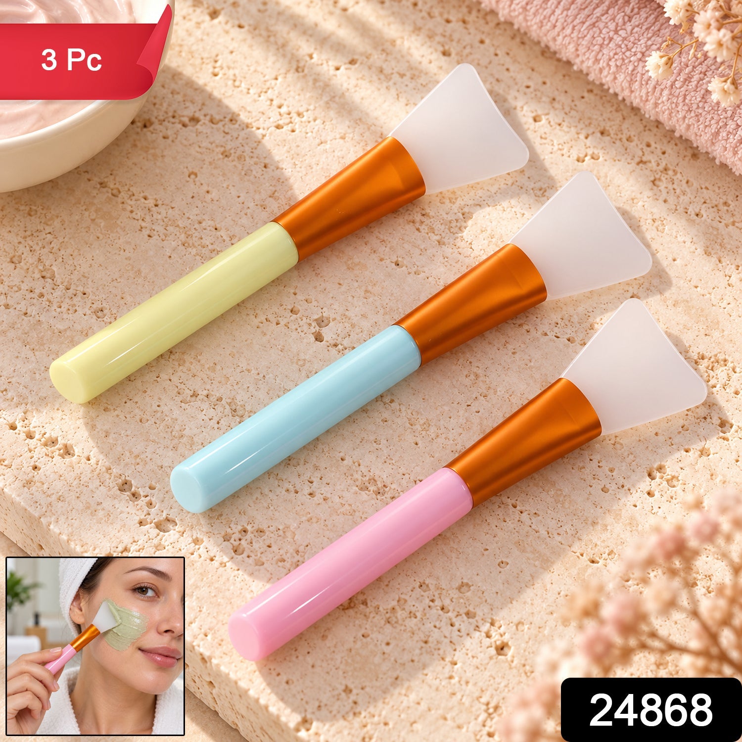 Three pastel-handled silicone face mask brushes on a stone surface, with a bowl of mask and a folded towel nearby.
