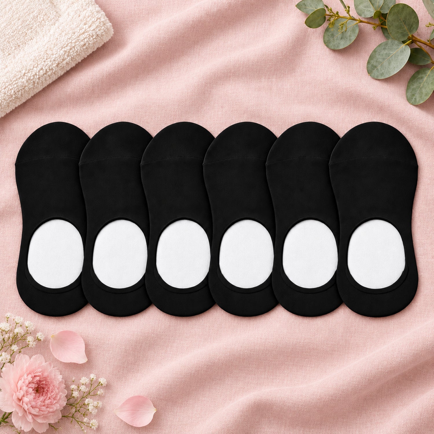 Six black low-cut socks with white heel grips, arranged on a pink fabric with flower petals.