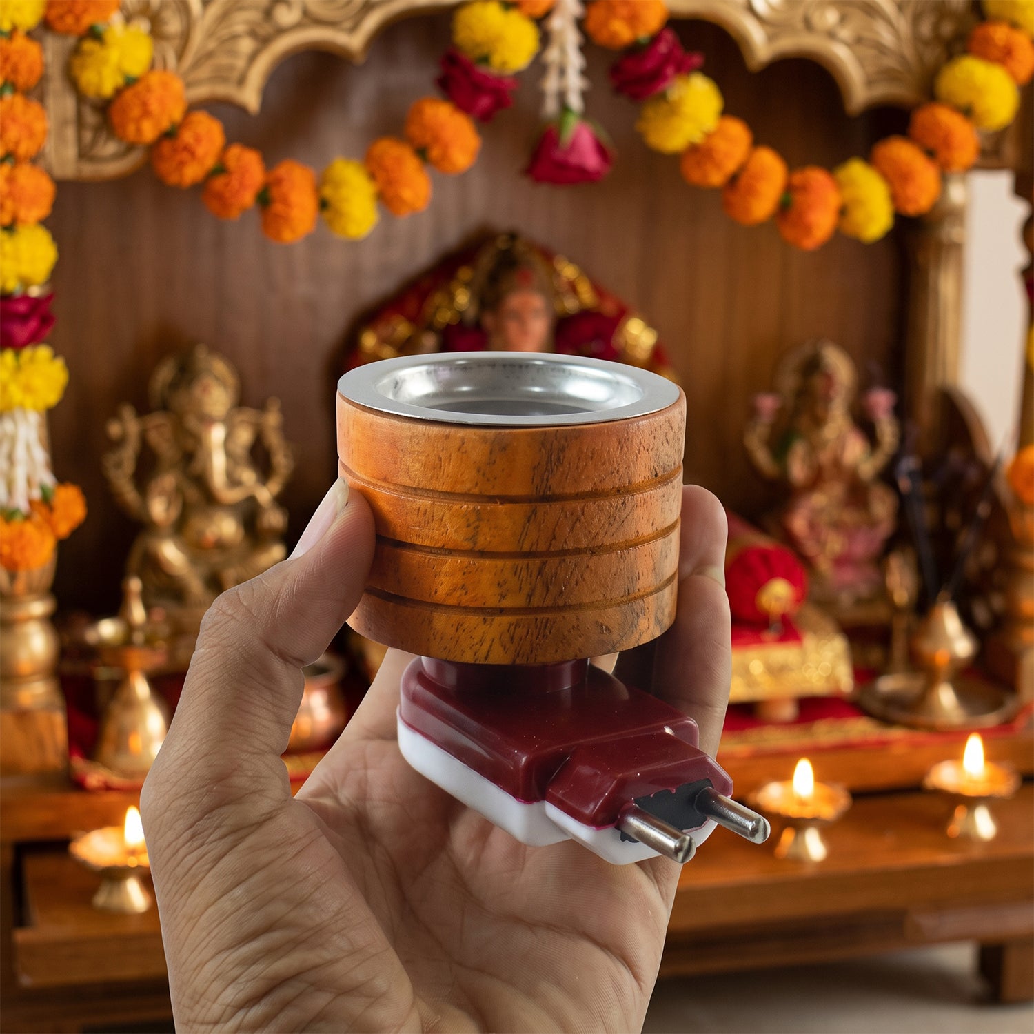 A hand holds a wooden electric camphor diffuser with a metal cup in front of a decorated shrine.