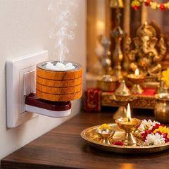 A wooden electric camphor diffuser plugged into a wall socket emits smoke, beside a lit lamp and flowers.