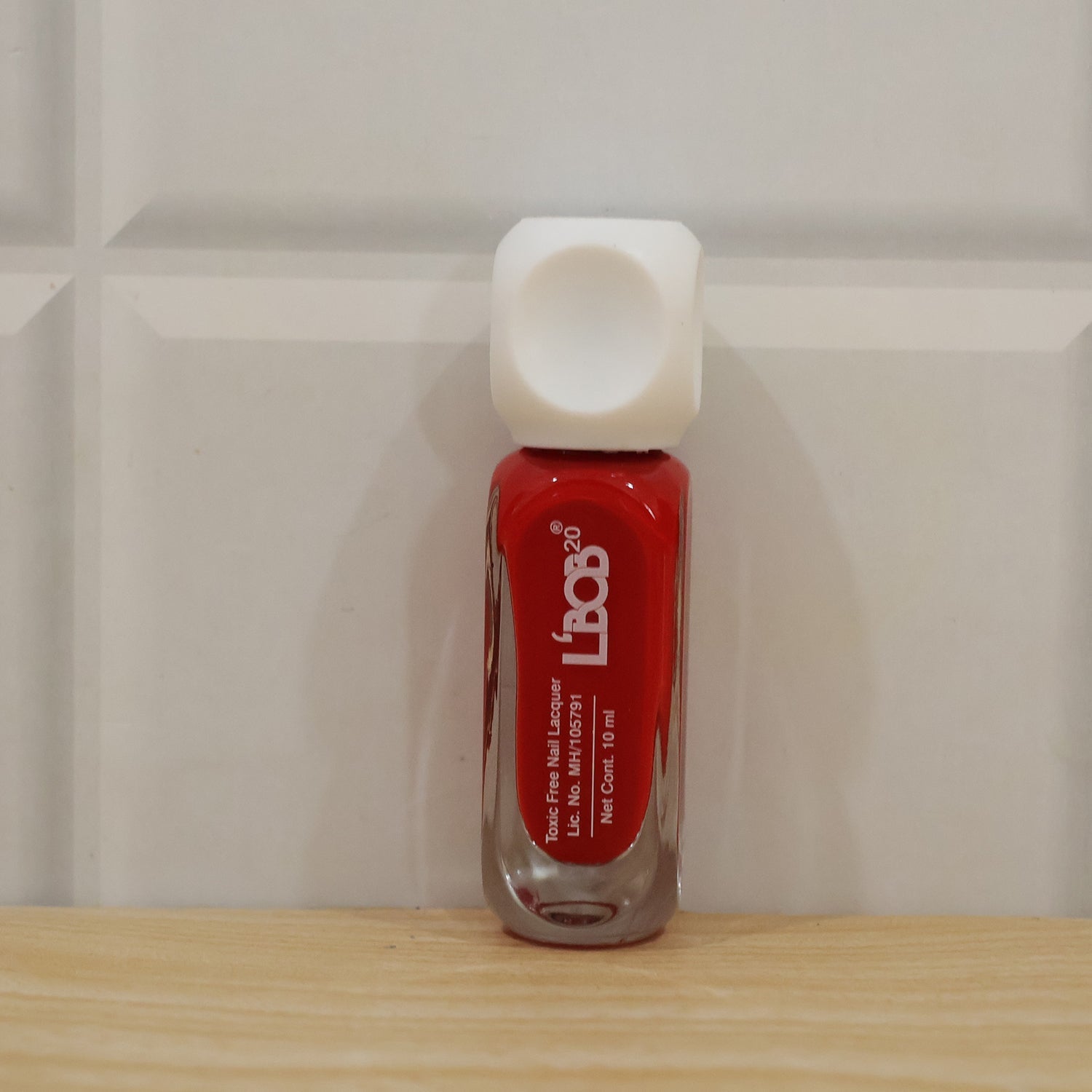 A red nail polish bottle with a white square cap stands on a wooden shelf against a white tiled wall.