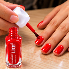A person applies red nail polish to their fingernails, with the bottle and brush visible on a wooden surface.