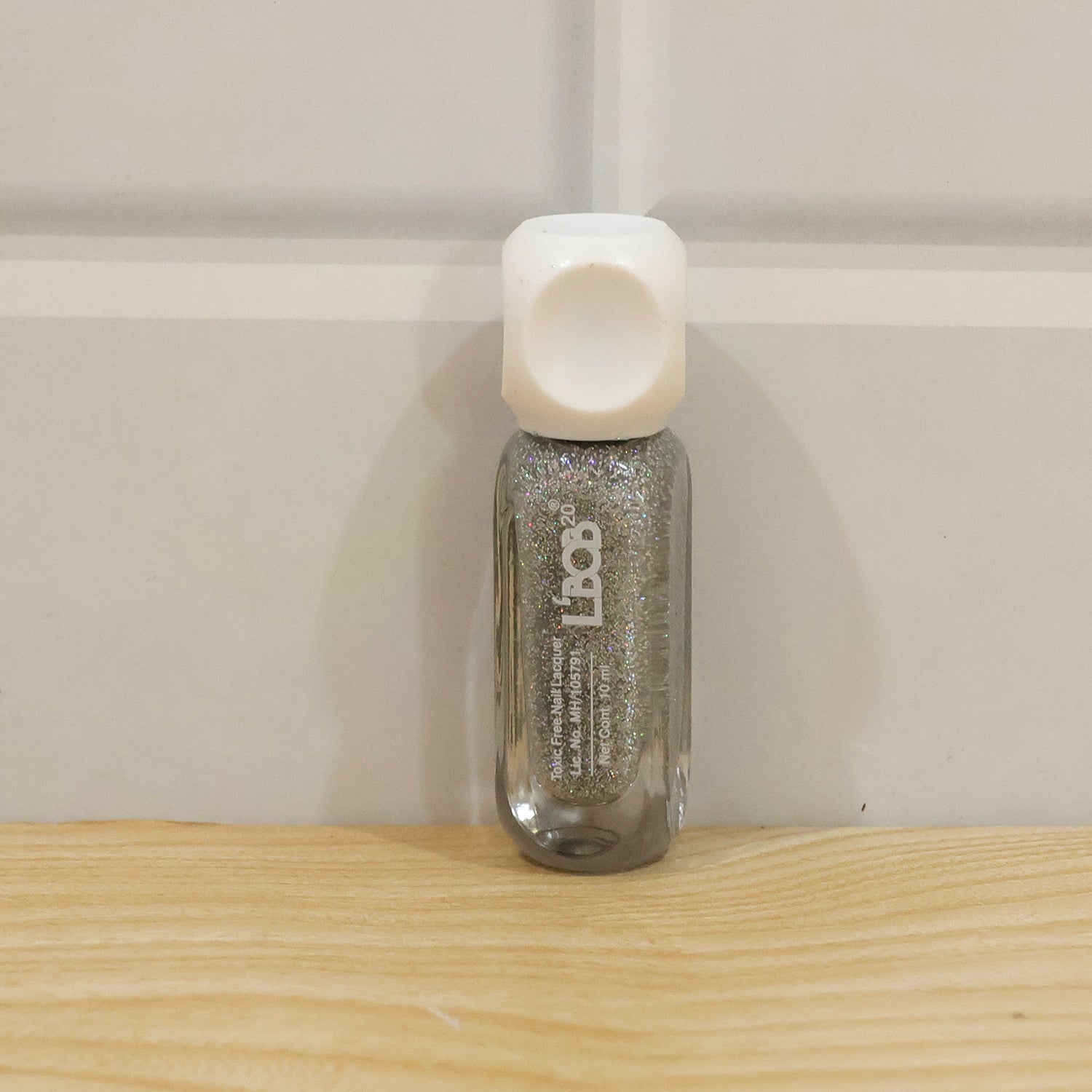 A small bottle of silver glitter nail polish with a white cap stands on a light wooden surface against a white wall.