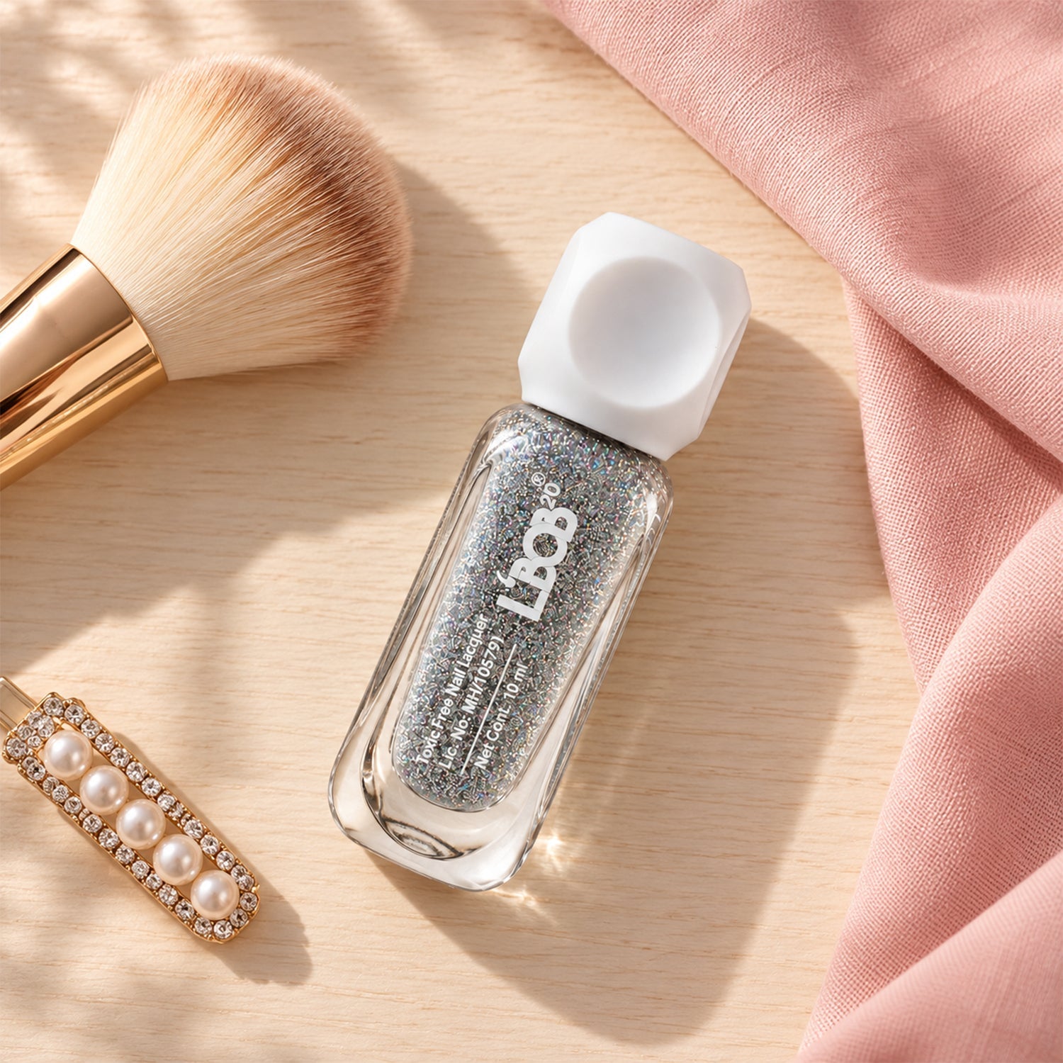 A clear bottle of silver glitter nail polish with a white cap rests on a wooden surface next to a makeup brush and pearl