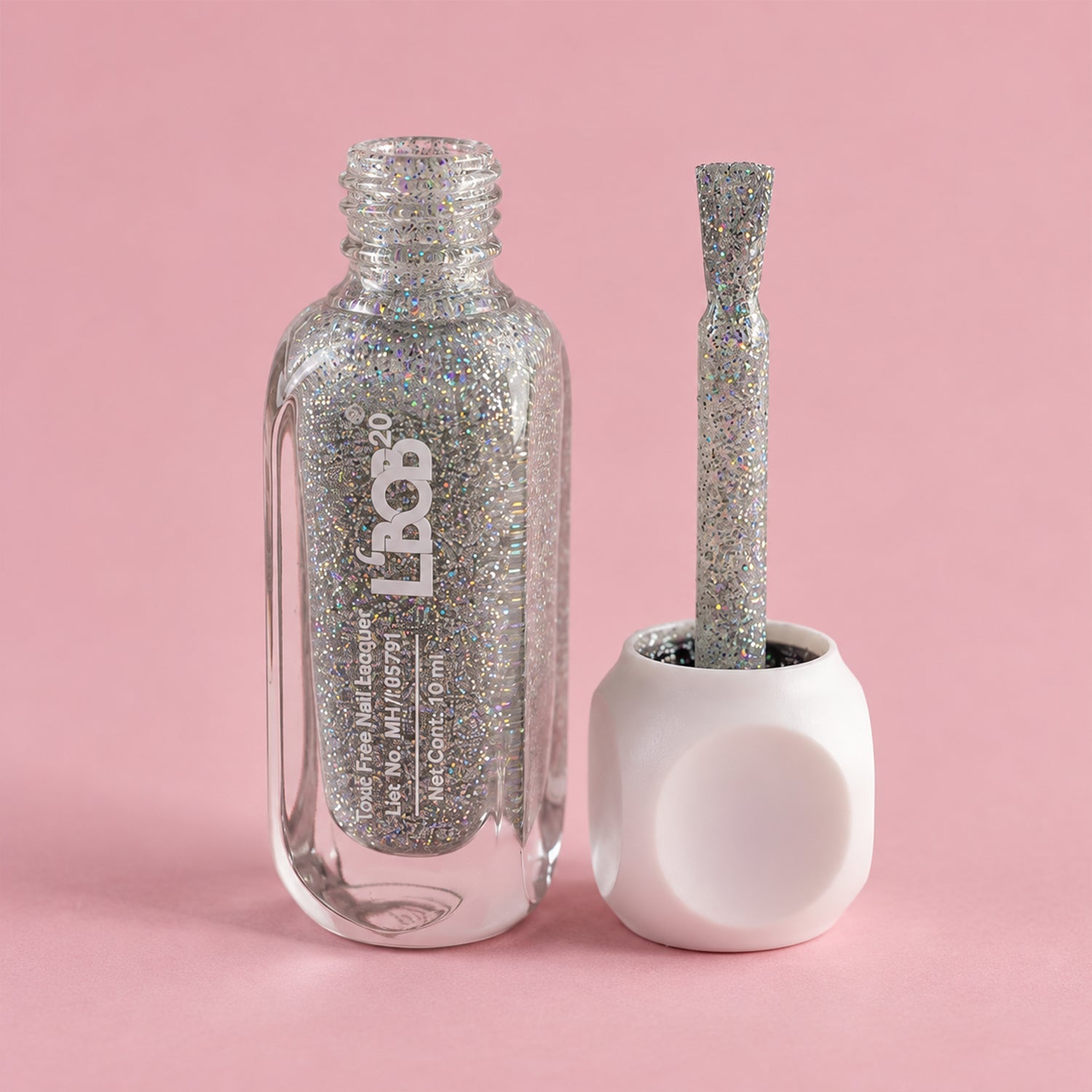 An open bottle of silver glitter nail polish sits next to its white cap with an attached brush on a pink background.