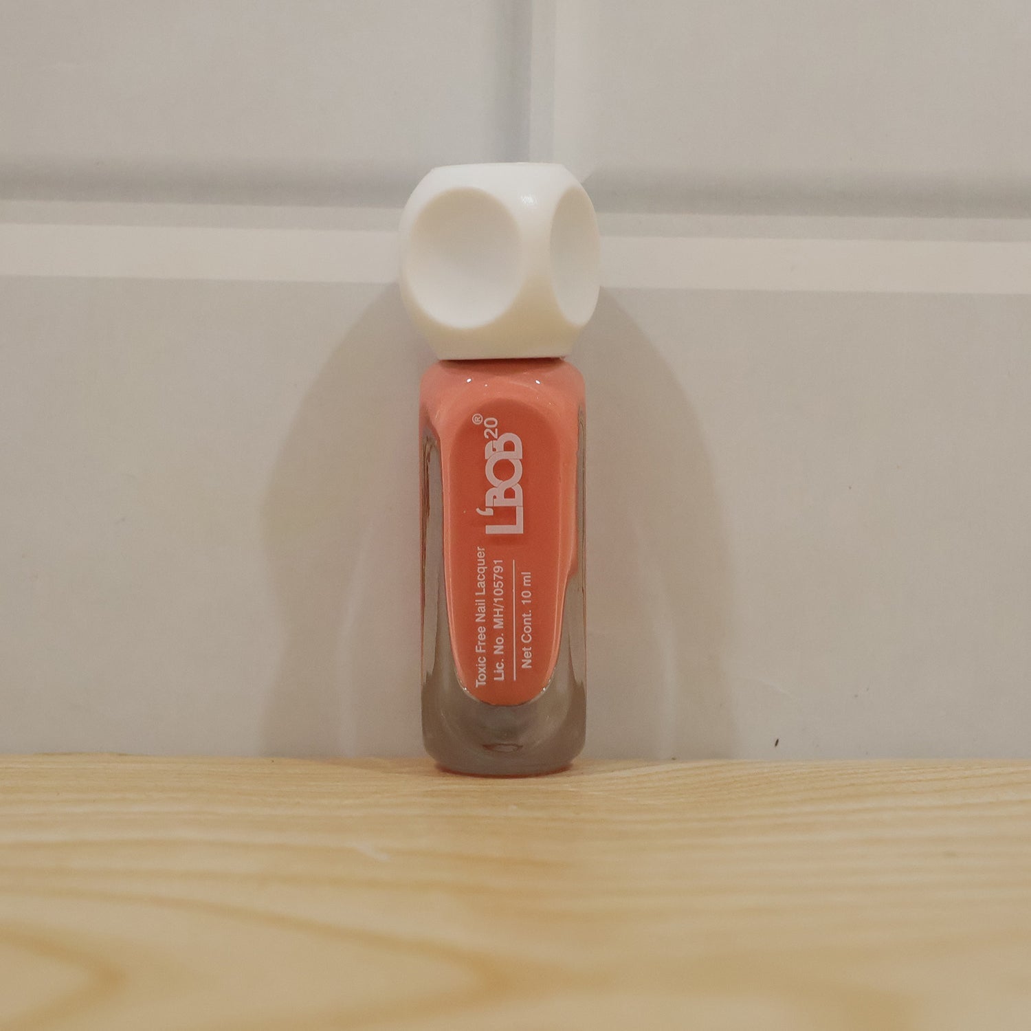 A single bottle of peach coral nail polish with a white dice-shaped cap stands on a light wooden surface against a white