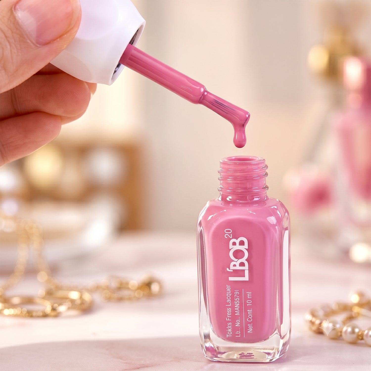 A hand holds a brush applicator dripping pink polish into an open bottle, surrounded by golden jewelry.