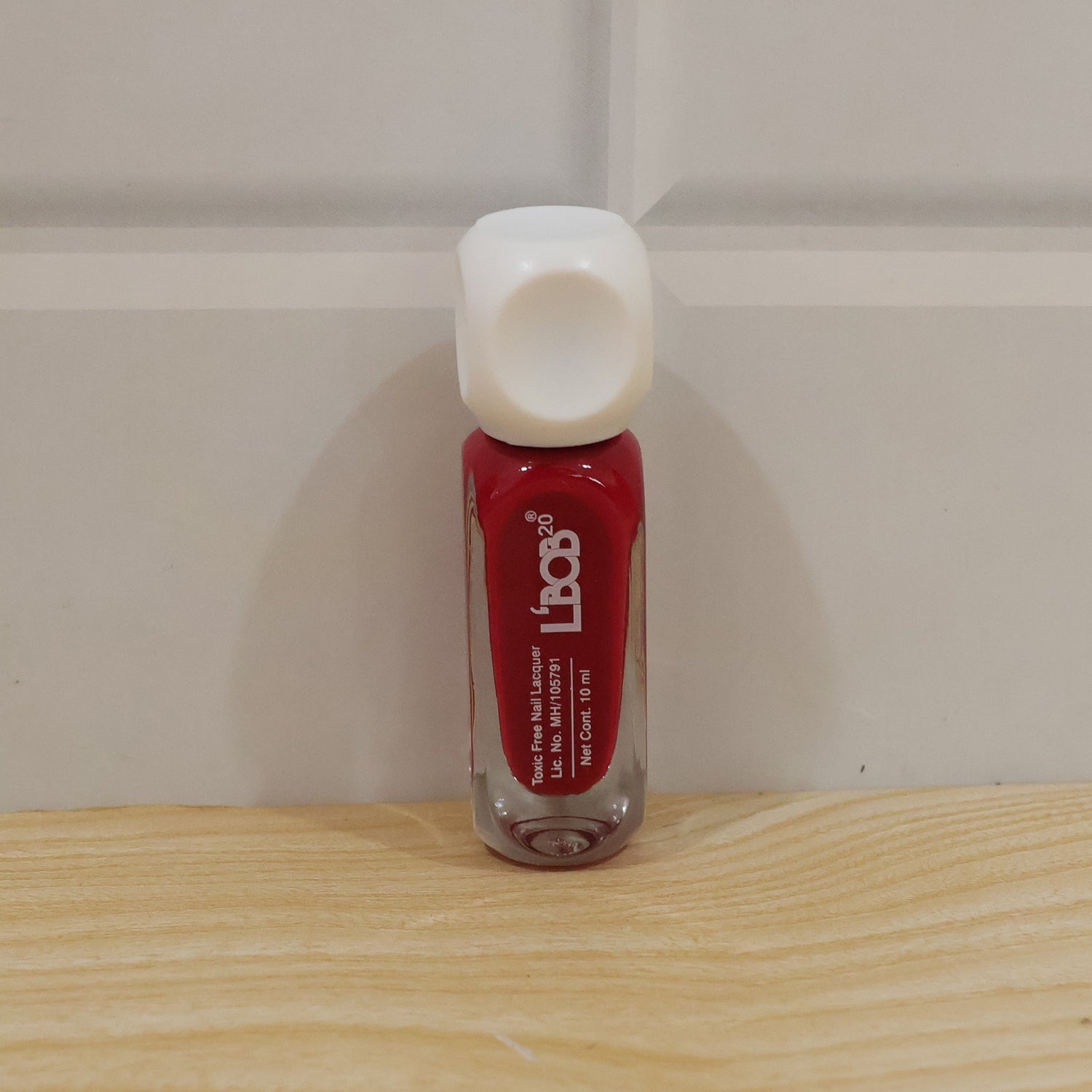 A bottle of red nail polish with a white cap stands on a light wooden surface against a white wall.