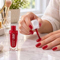 A person applies bright red nail polish to their fingernails with a brush, next to an open bottle.
