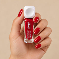 A hand holds a small clear bottle of red nail polish with a white square cap, against a light background.