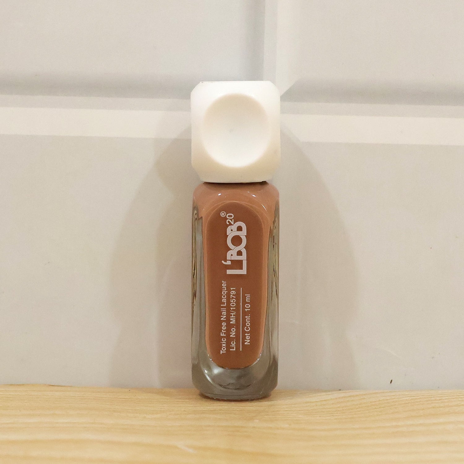 A small bottle of nude brown nail polish with a white cap stands on a wooden surface against a white door.