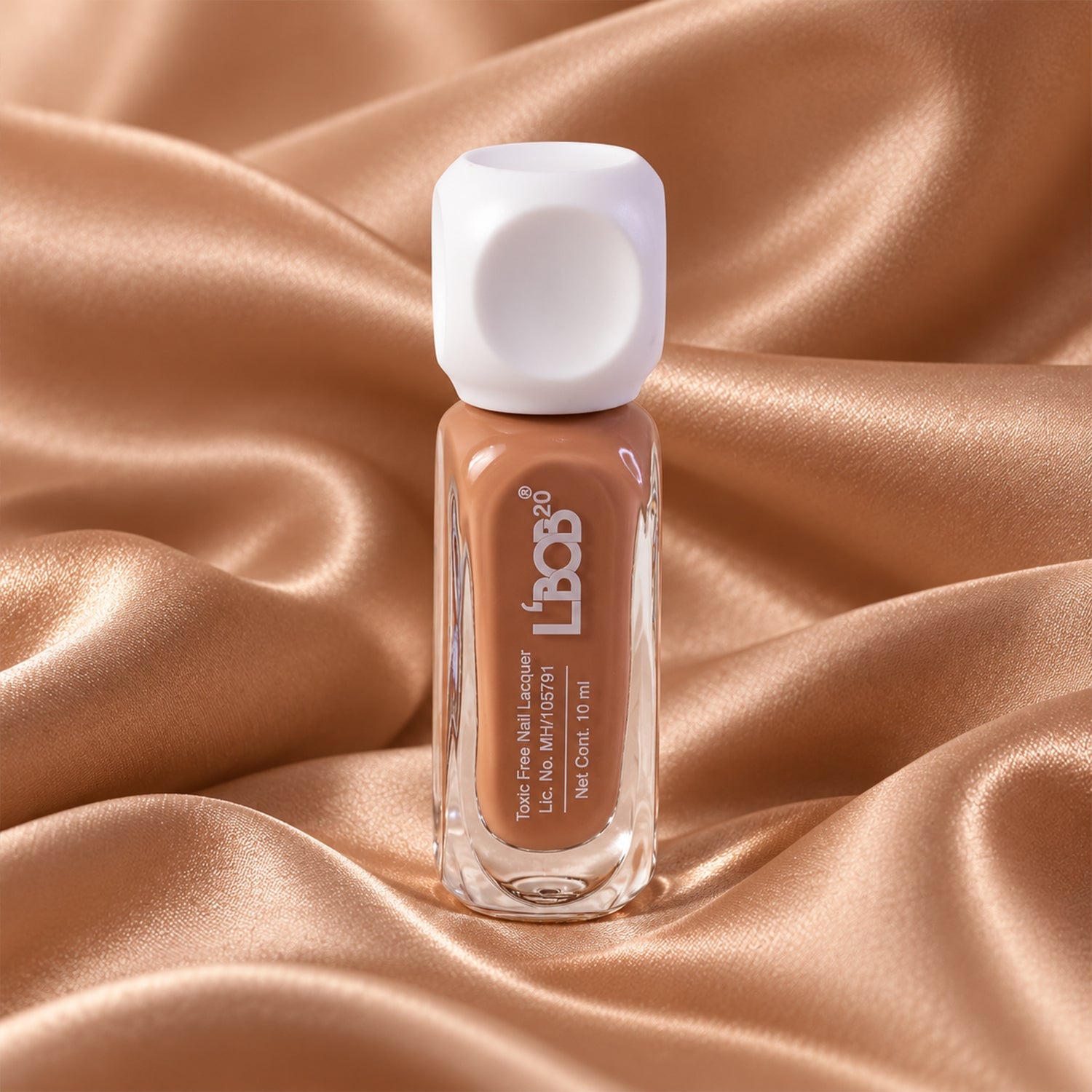 A small bottle of nude brown nail polish with a white cap is centered on a draped golden satin fabric.