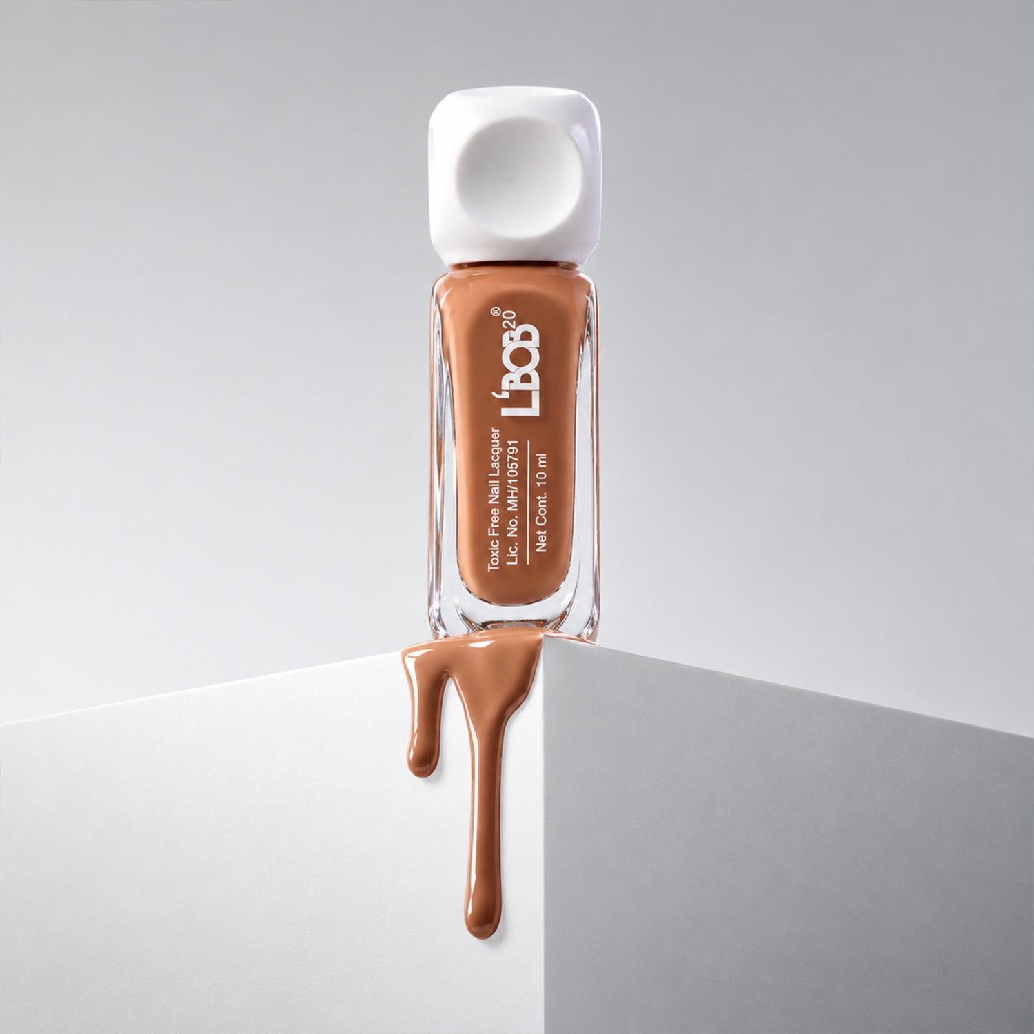 A bottle of nude brown nail polish with a white cap sits on a white block with polish dripping down.