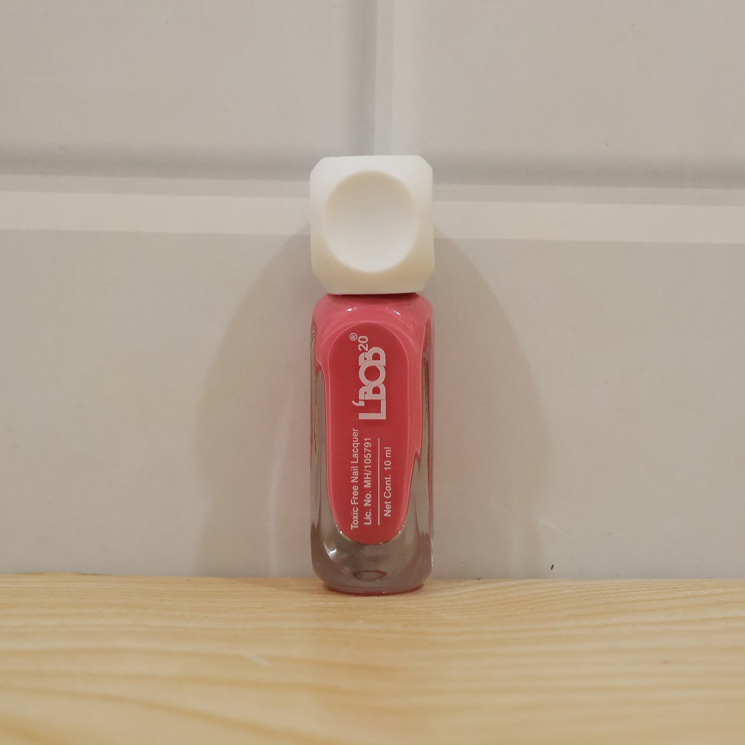 A pink nail polish bottle with a white cap stands upright on a light wooden surface against a white paneled wall.