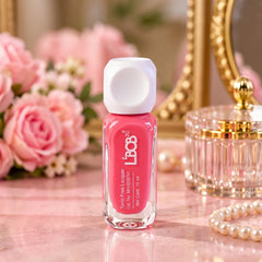 A vibrant pink nail polish bottle with a white square cap stands on a marble surface, surrounded by roses and pearls.