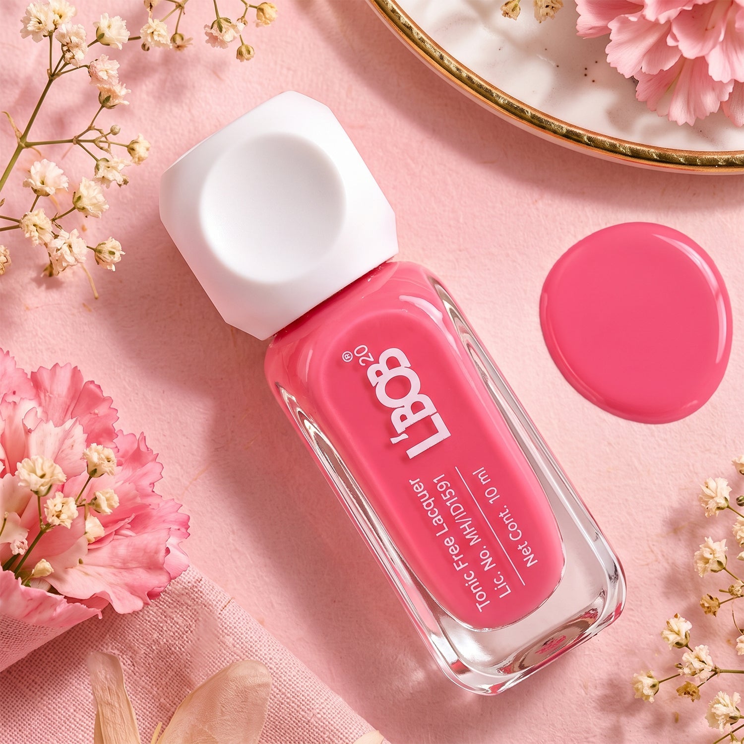 A pink nail polish bottle with a white cap lies on a pink surface next to a dollop of matching polish and small flowers.