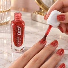 A person applies red glitter nail polish from an open bottle to their fingernails.