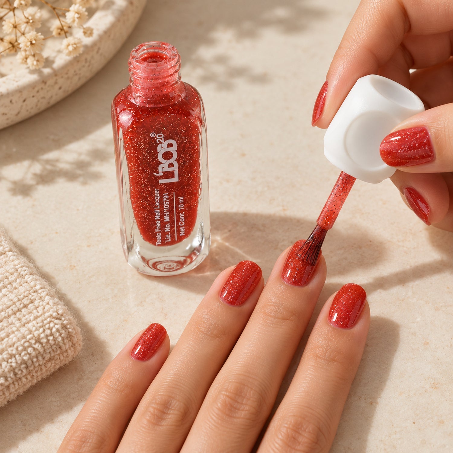A person applies red glitter nail polish to their fingernails, with the open bottle beside a white towel.