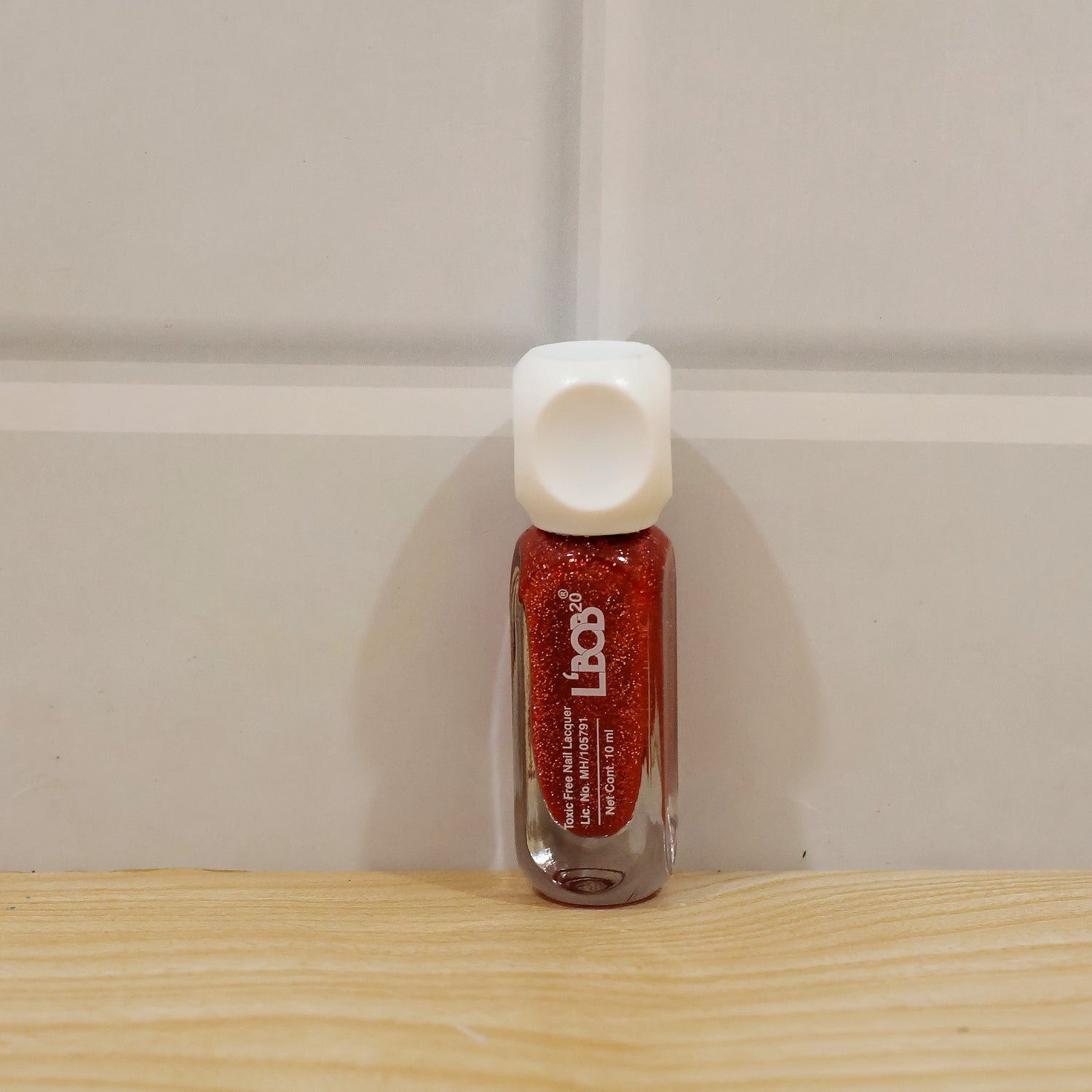 A small, clear glass bottle of red glitter nail polish with a white cap stands on a wooden surface.