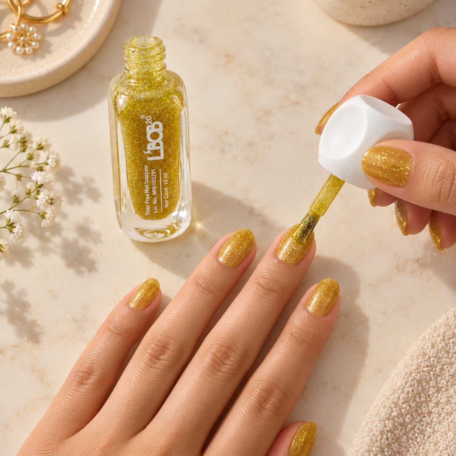 A person applies gold glitter nail polish to their fingernails, with a bottle of polish and a white towel nearby.
