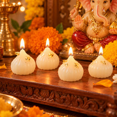 Four lit modak-shaped white wax candles with gold flakes on a wooden surface, with a Ganesha idol in the background.