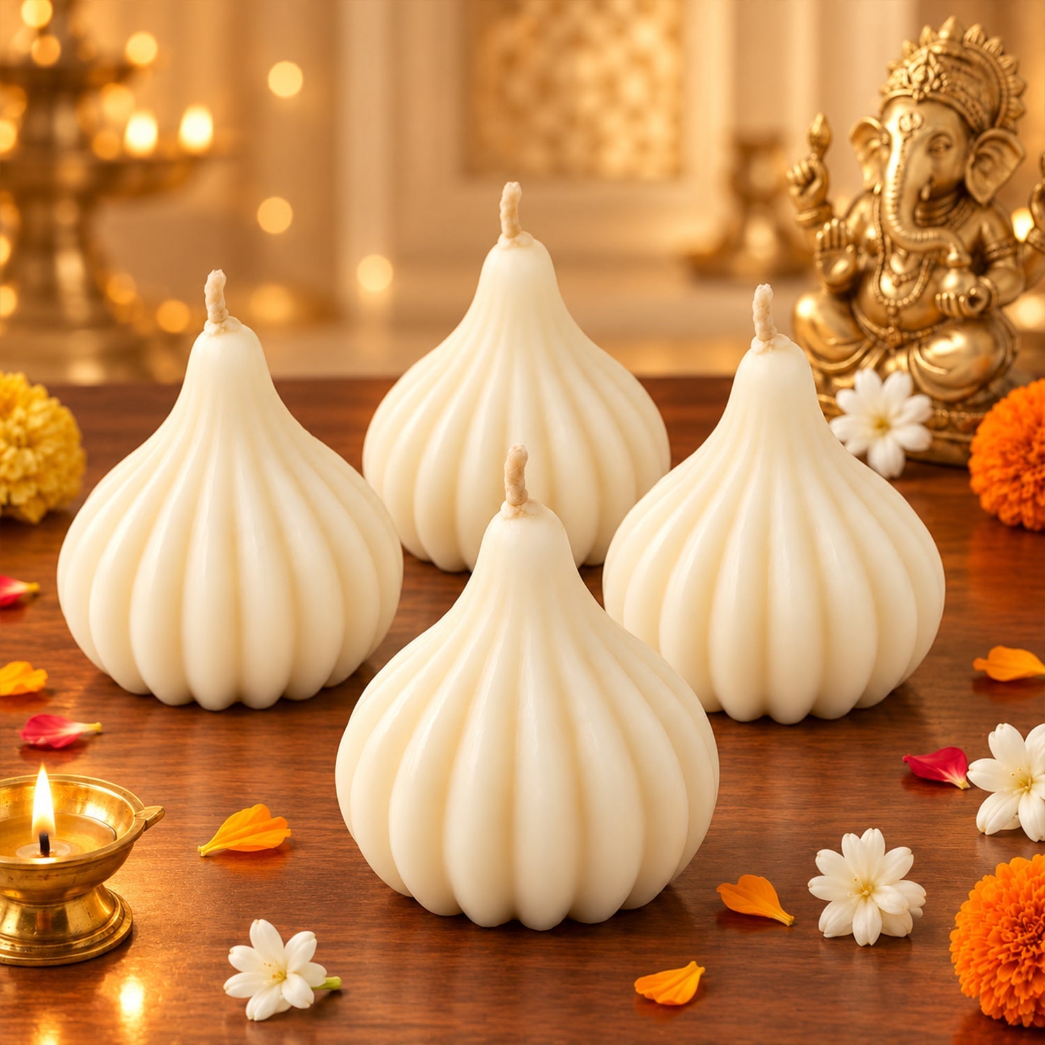 Four unlit white modak-shaped candles arranged on a wooden surface, with scattered flower petals and a Ganesha idol in the