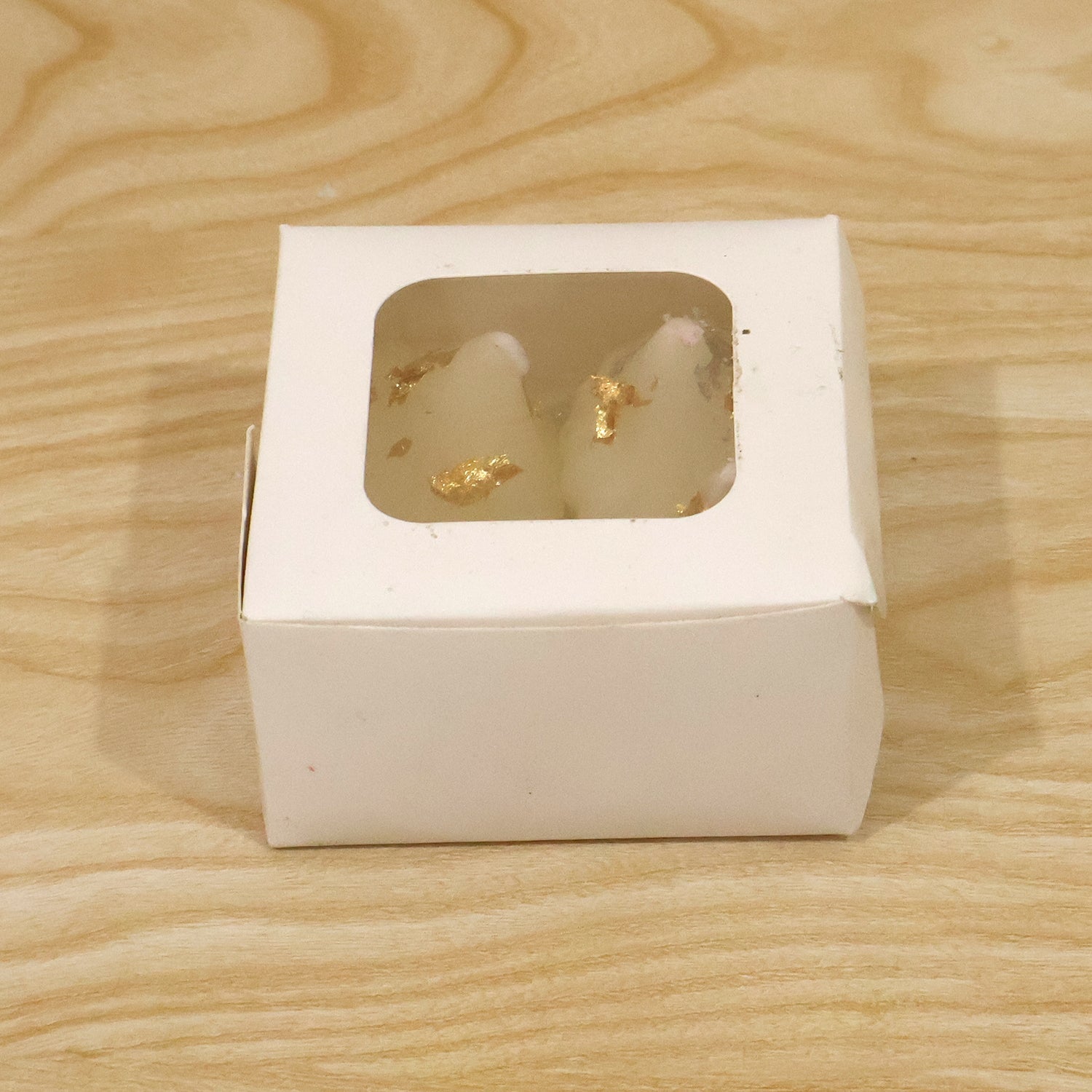 A small white cardboard box with a clear window, displaying two modak-shaped white candles with gold flakes inside.