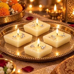 Four lit diamond-shaped cream candles with silver foil accents on a gold ornate tray, surrounded by warm glowing lights and