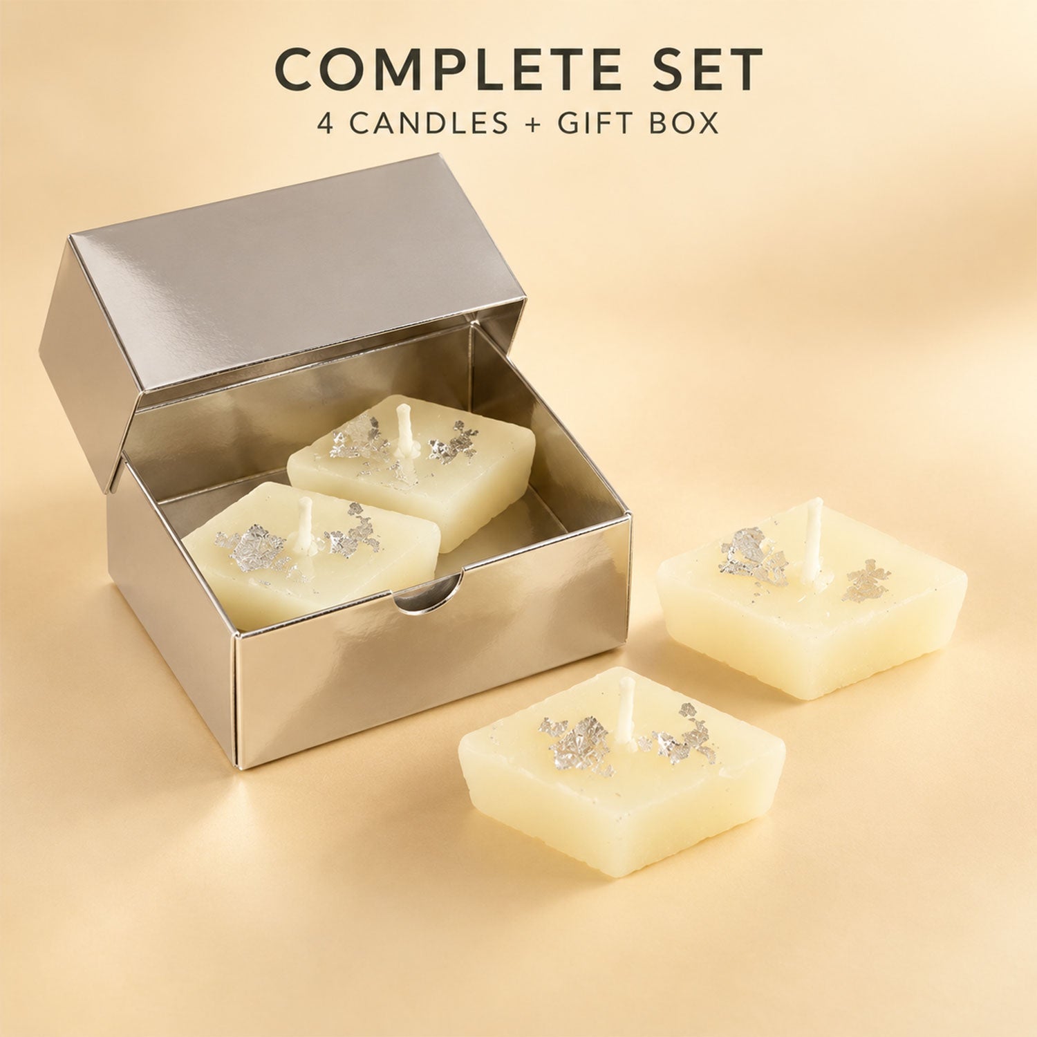 Two diamond-shaped cream candles with silver foil inside an open silver gift box, with two more candles placed outside the