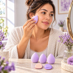 A woman applies makeup with a lavender blending sponge, with three sponges and two puffs on a table.