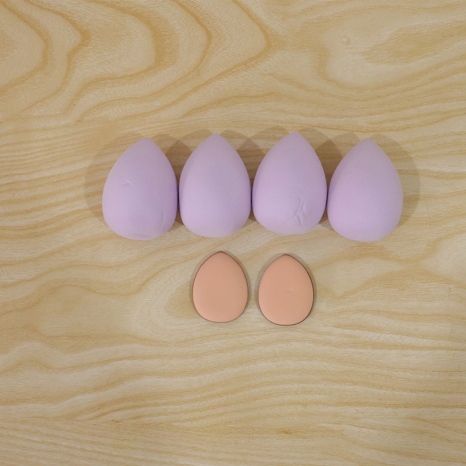 Four lavender teardrop sponges and two peach oval puffs are arranged on a light wooden surface.