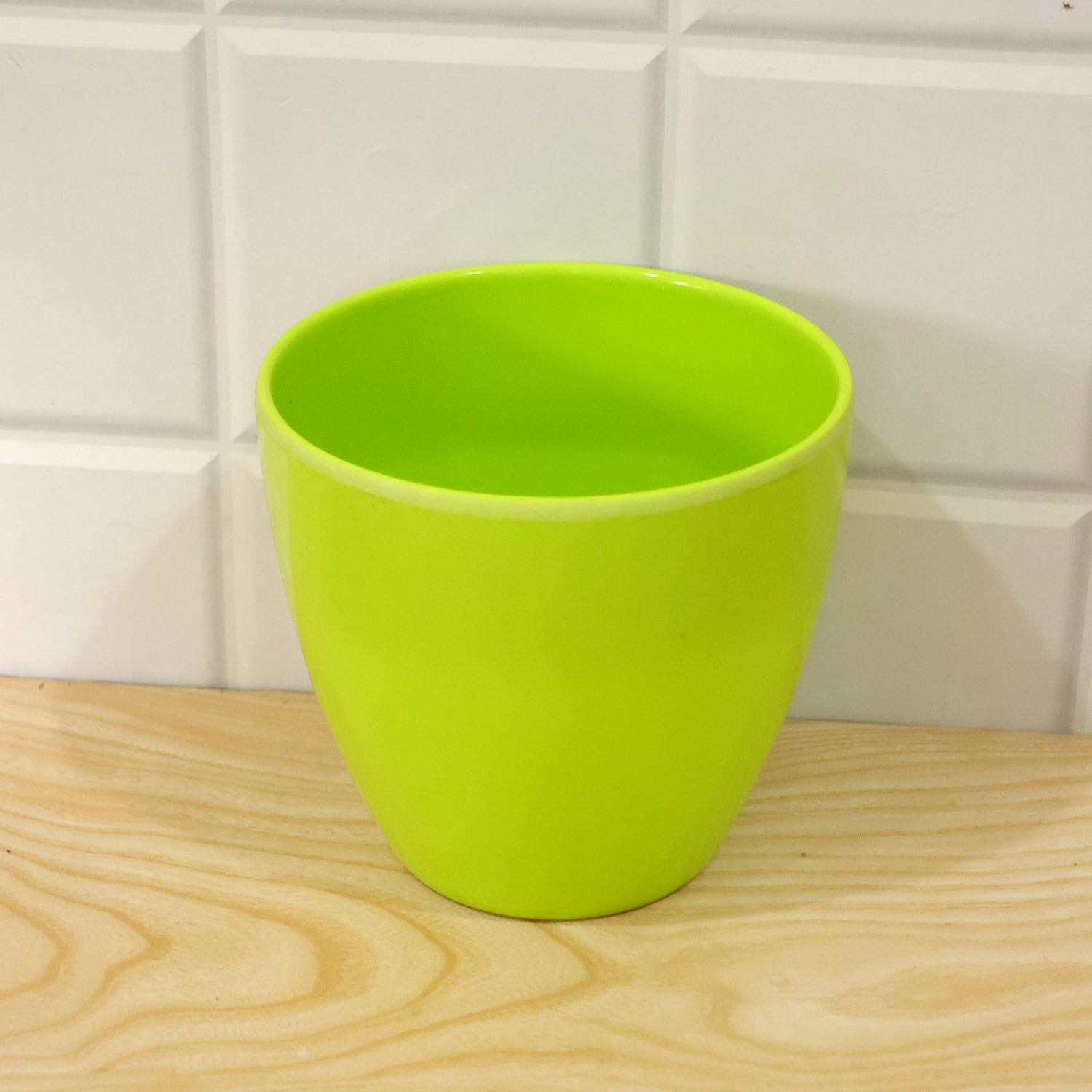 An empty, lime green plastic pot resting on a light wood surface, with white tiled wall in the background.