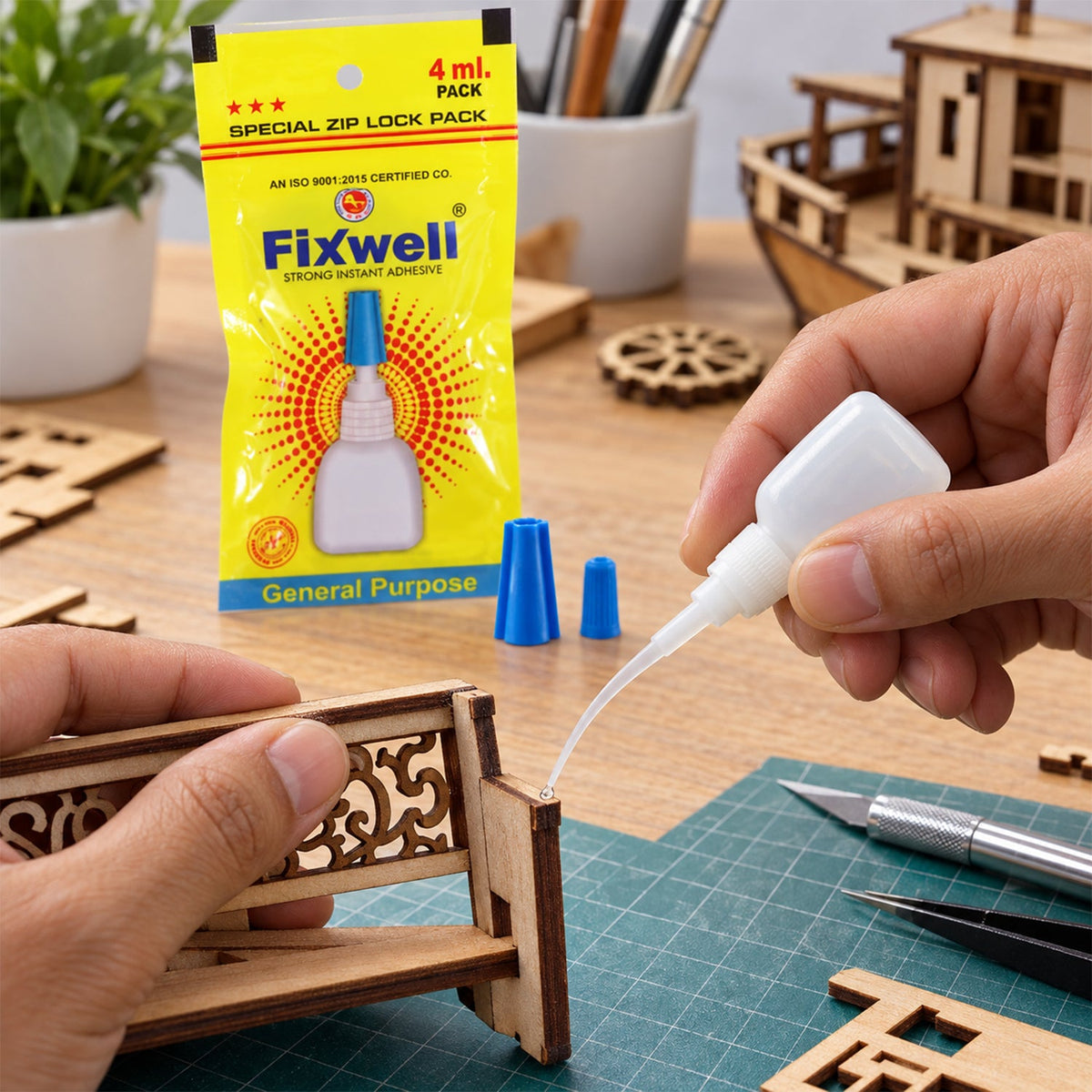 A person applies clear glue from a small white bottle with a fine nozzle to a wooden miniature structure. A yellow packet of
