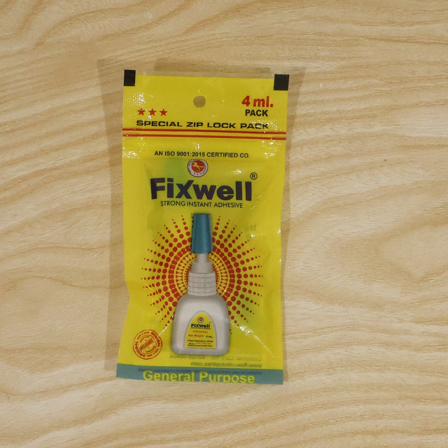 A yellow Fixwell glue packet with a blue and white glue bottle design is centered on a light wooden background.