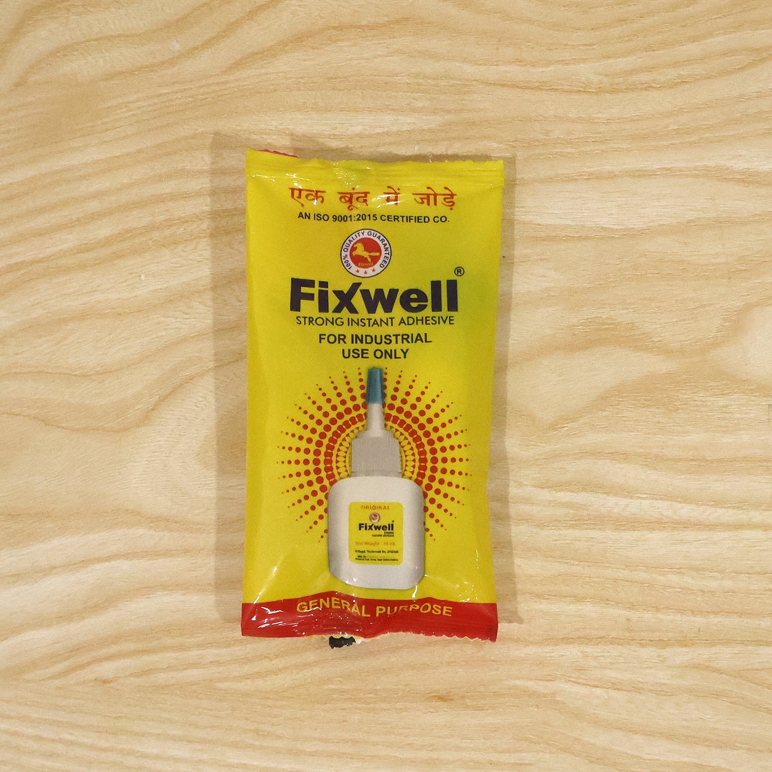A yellow Fixwell instant adhesive packet with a red stripe is centered on a light wooden surface.