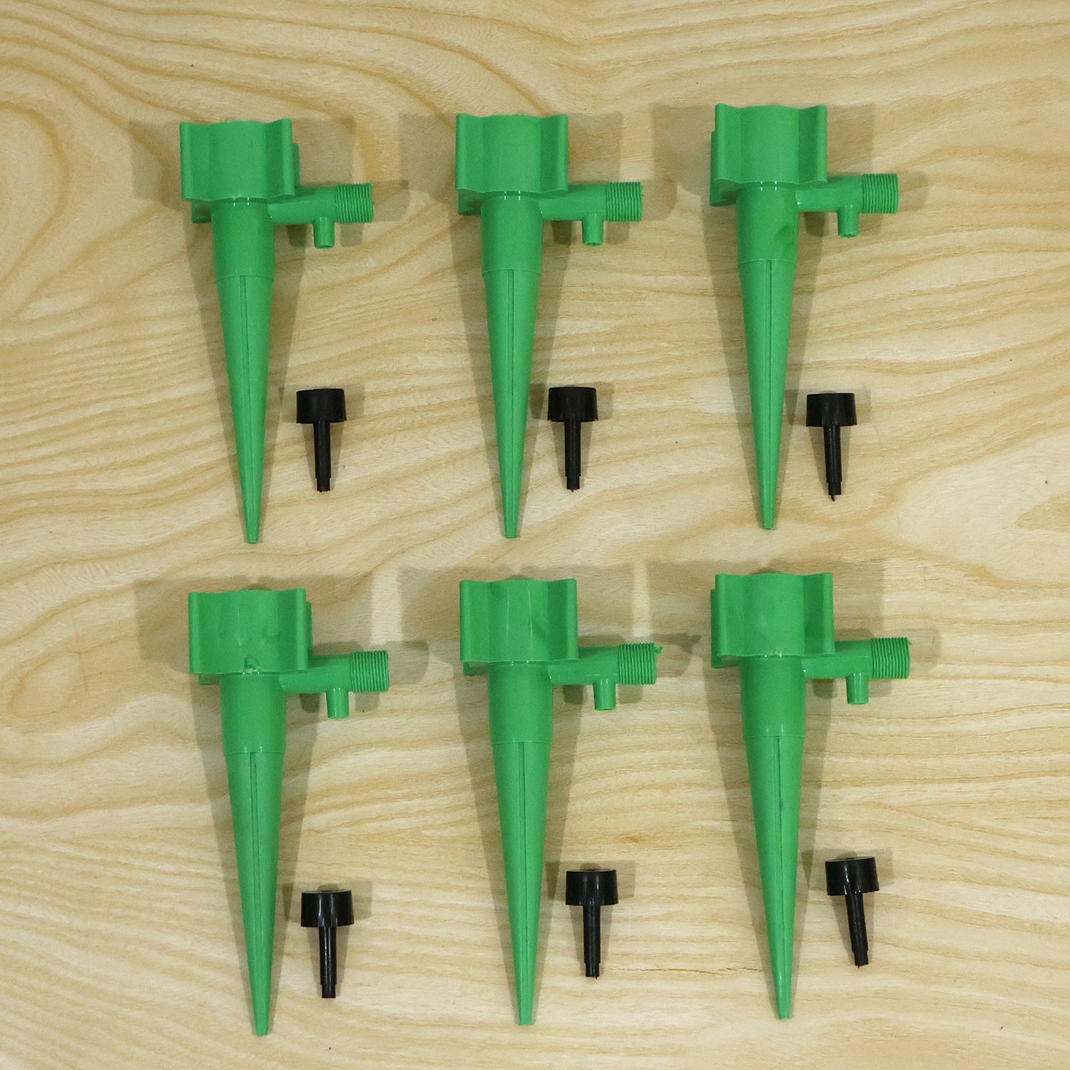 Six green plastic plant watering spikes and six small black caps are arranged on a light wooden surface.