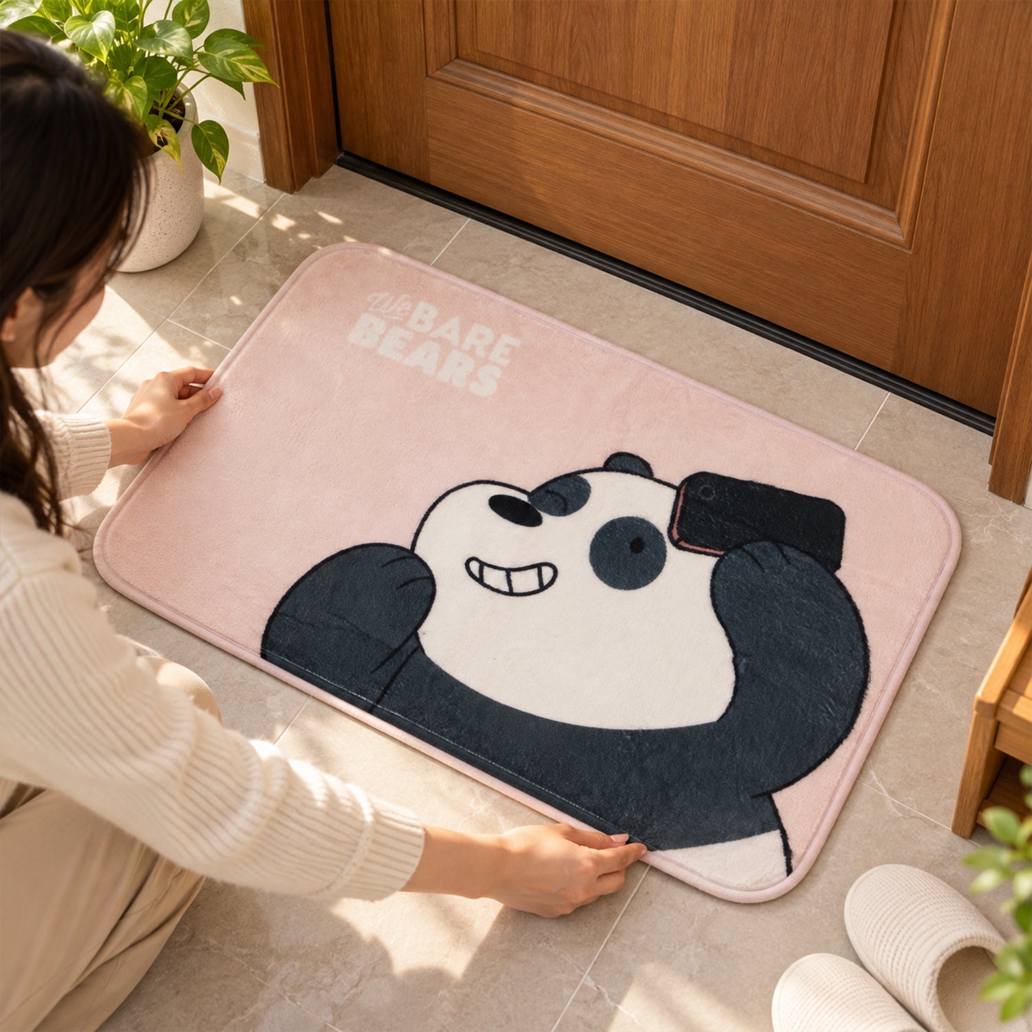 A person kneels on a wooden floor, adjusting a pink panda mat near a door and slippers.