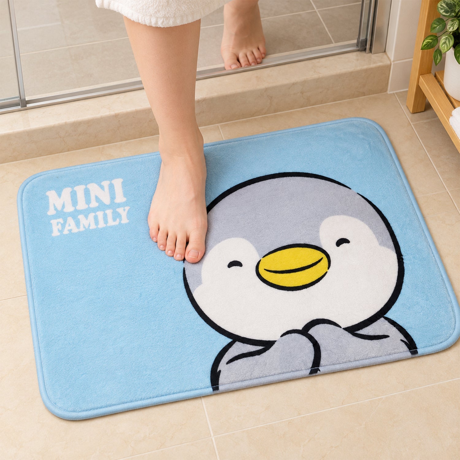A person's bare feet are shown on a blue bath mat with a smiling penguin graphic, located in a tiled bathroom.
