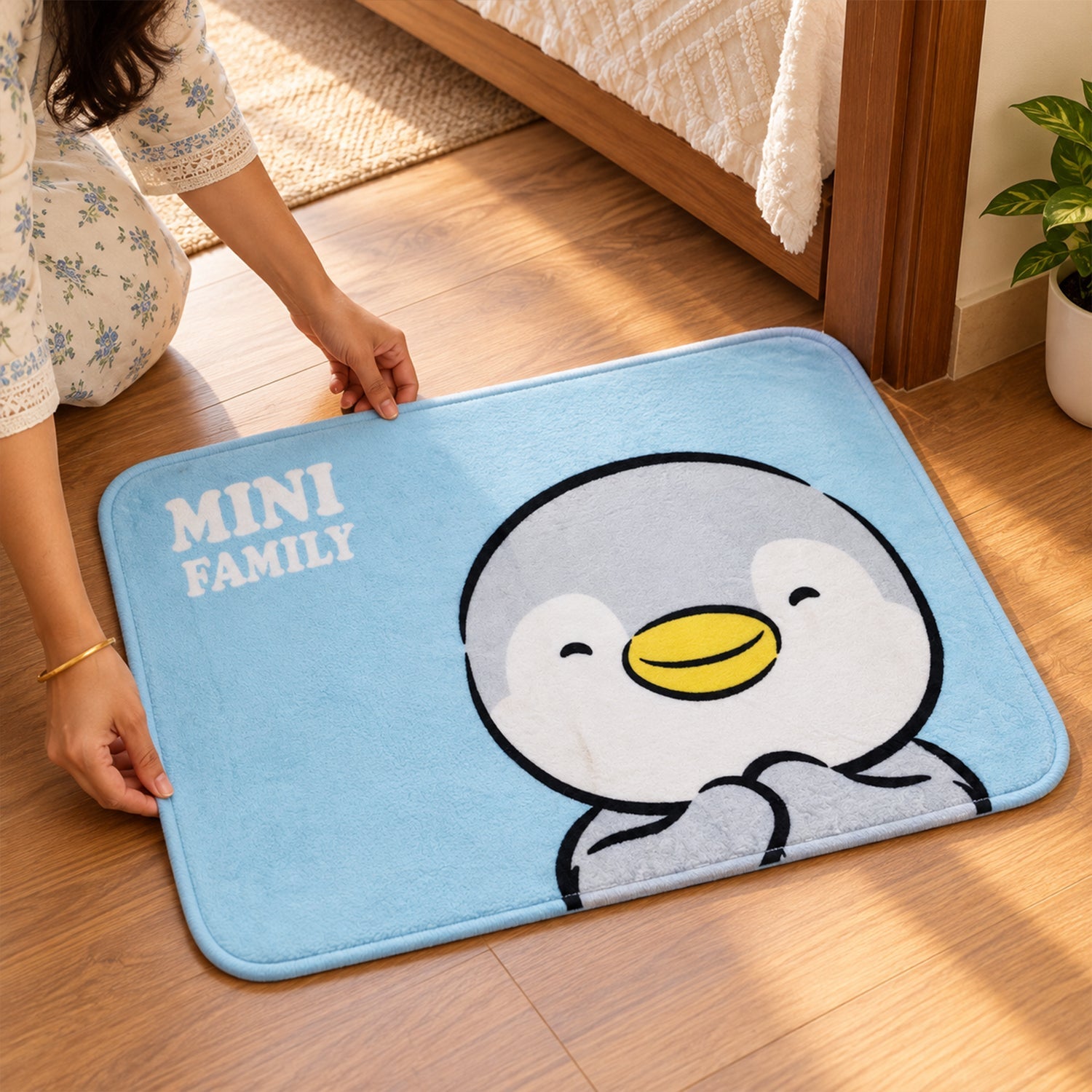 A person's hands adjust a blue bath mat featuring a cartoon penguin design on a wooden floor beside a bed.