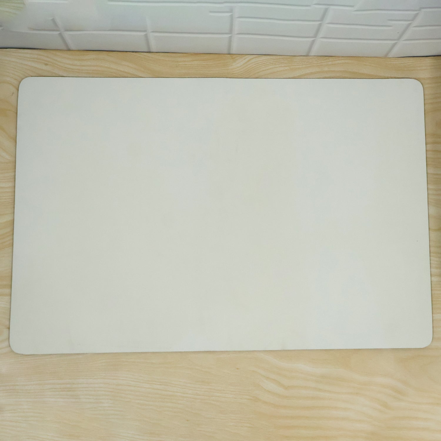 A cream rectangular mat with rounded corners lies flat on a light wood floor, against a white tiled wall.