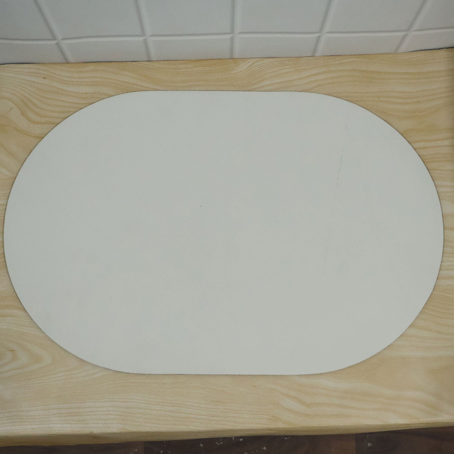 A plain white oval mat resting on a light wooden surface with white tiled background.